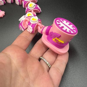 May include: A pink and white articulated toy, featuring a top hat and teacup design. The top hat has a clock face and the number 10/6. The toy is held in a hand, showcasing its flexible, connected segments.