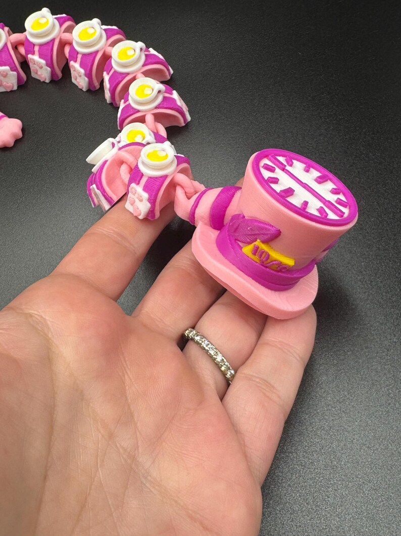 May include: A pink and white articulated toy, featuring a top hat and teacup design. The top hat has a clock face and the number 10/6. The toy is held in a hand, showcasing its flexible, connected segments.