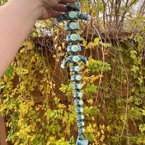 May include: A teal and grey rain chain with a unique, articulated design. The chain features a series of connected, circular elements with green accents, resembling a stylized figure. The rain chain is suspended against a backdrop of autumn foliage.