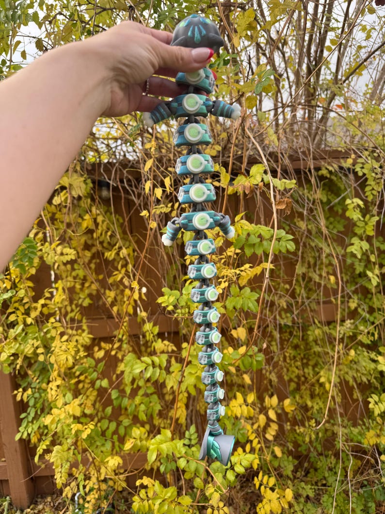 May include: A teal and grey rain chain with a unique, articulated design. The chain features a series of connected, circular elements with green accents, resembling a stylized figure. The rain chain is suspended against a backdrop of autumn foliage.