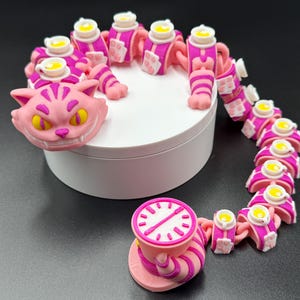 May include: A collection of pink and white figurines inspired by the Cheshire Cat. The set features a cat's head, a teacup with a clock face, and a chain of body segments, each with a teacup. The figurines are displayed on a white platform.