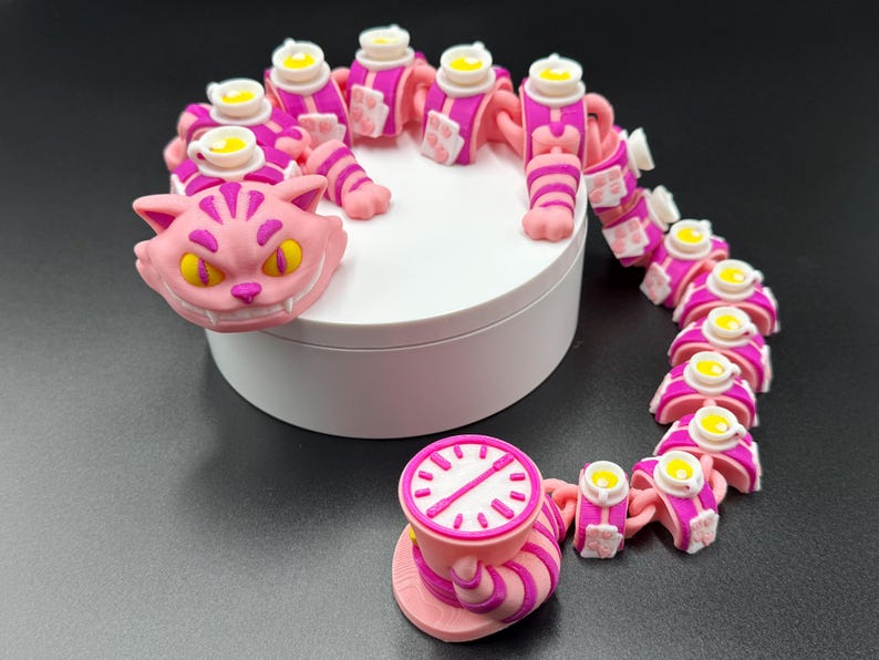 May include: A collection of pink and white figurines inspired by the Cheshire Cat. The set features a cat's head, a teacup with a clock face, and a chain of body segments, each with a teacup. The figurines are displayed on a white platform.