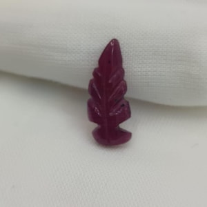 Könnte beinhalten: Ein kleiner, geschnitzter Edelstein in Form eines Blattes. Der Edelstein hat eine tiefe, satte burgunderrote Farbe. Das Blattdesign hat detaillierte Adern und eine abgerundete Basis. Der Edelstein ist vor einem weißen Hintergrund platziert.
