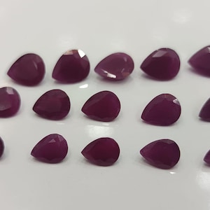Könnte beinhalten: Fünfzehn facettierte, tropfenförmige Edelsteine in einem tiefen, satten Burgunderrot. Die Edelsteine sind auf einer weißen Oberfläche angeordnet und weisen jeweils eine polierte, reflektierende Oberfläche auf. Die Edelsteine sind wahrscheinlich Rubine.