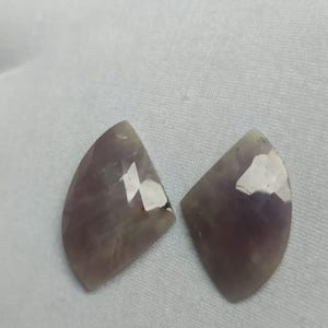 Könnte beinhalten: Zwei Amethyst-Edelsteine, jeweils mit einer facettierten, halbkreisförmigen Form. Die Steine zeigen eine Reihe von Lilatönen und sind vor einem weißen Hintergrund platziert. Die Edelsteine sind wahrscheinlich für die Schmuckherstellung oder das Sammeln bestimmt.