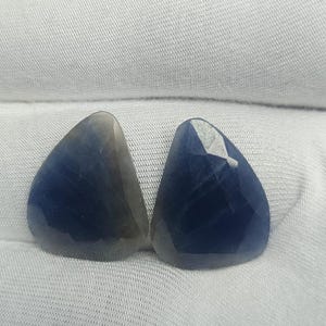 Könnte beinhalten: Zwei facettierte, dreieckige, saphirblaue Edelsteine. Die Edelsteine werden auf einem weißen, strukturierten Stoff präsentiert. Die Steine haben eine polierte, reflektierende Oberfläche und eine tiefblaue Farbe, wobei einige Bereiche heller erscheinen.