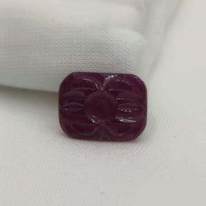 Könnte beinhalten: Ein rechteckiger, tiefroter Edelstein mit geschnitztem Blumendesign. Der Edelstein ist vor einem weißen Hintergrund platziert. Das geschnitzte Design zeigt eine zentrale Blume mit blütenblattartigen Formen.