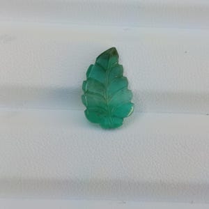 Könnte beinhalten: Ein geschnitzter grüner Edelstein in Blattform mit detaillierten Adern. Der Edelstein hat einen leuchtenden Grünton und ist vor einem weißen Hintergrund platziert. Das Blattdesign ist filigran und gut definiert und zeigt die Handwerkskunst der Schnitzerei.