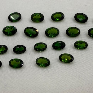 Può includere: Una collezione di gemme verdi di forma ovale. Le gemme sono sparse su una superficie bianca, mostrando le loro sfaccettature lucide e il loro colore intenso. Ideale per la creazione di gioielli.