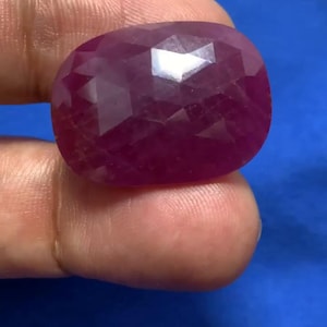 Könnte beinhalten: Ein facettierter, oval geformter Rubin-Edelstein. Der Edelstein hat eine tiefe, satte rote Farbe mit mehreren Facetten, die das Licht reflektieren. Der Rubin wird vor einem blauen Hintergrund gehalten.