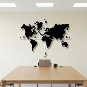Puede incluir: Un mapa del mundo de metal negro con un diseño de brújula. El mapa está montado en una pared blanca en un entorno de oficina moderno. Una mesa de madera, sillas y un ordenador portátil están en primer plano.