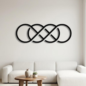 Può includere: Arte da parete in metallo nero con un design astratto di quattro cerchi interconnessi. L'opera d'arte è montata su una parete bianca sopra un divano bianco e un tavolino di legno. Il design aggiunge un tocco moderno al soggiorno.
