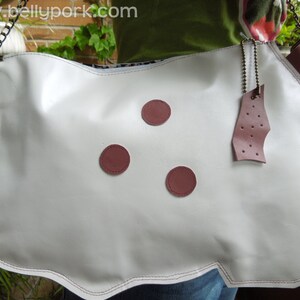 May include: White leather handbag with a chain strap and three burgundy colored buttons. The bag has a scalloped edge and a burgundy leather tag with holes.