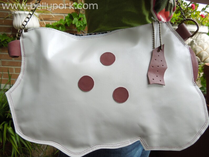 May include: White leather handbag with a chain strap and three burgundy colored buttons. The bag has a scalloped edge and a burgundy leather tag with holes.