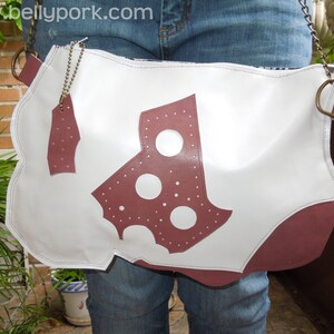 May include: A white leather bag with a burgundy leather design. The bag has a strap and a metal buckle. The bag is worn over the shoulder.