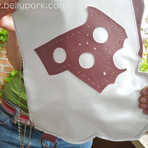 May include: A white leather bag with a burgundy leather patch featuring a cut-out design with three circles. The bag has a silver chain strap.