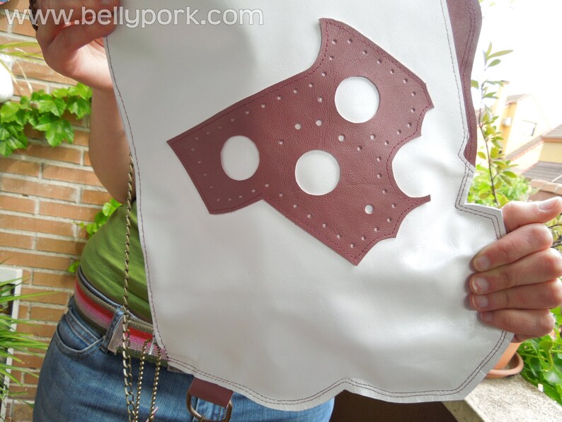 May include: A white leather bag with a burgundy leather patch featuring a cut-out design with three circles. The bag has a silver chain strap.