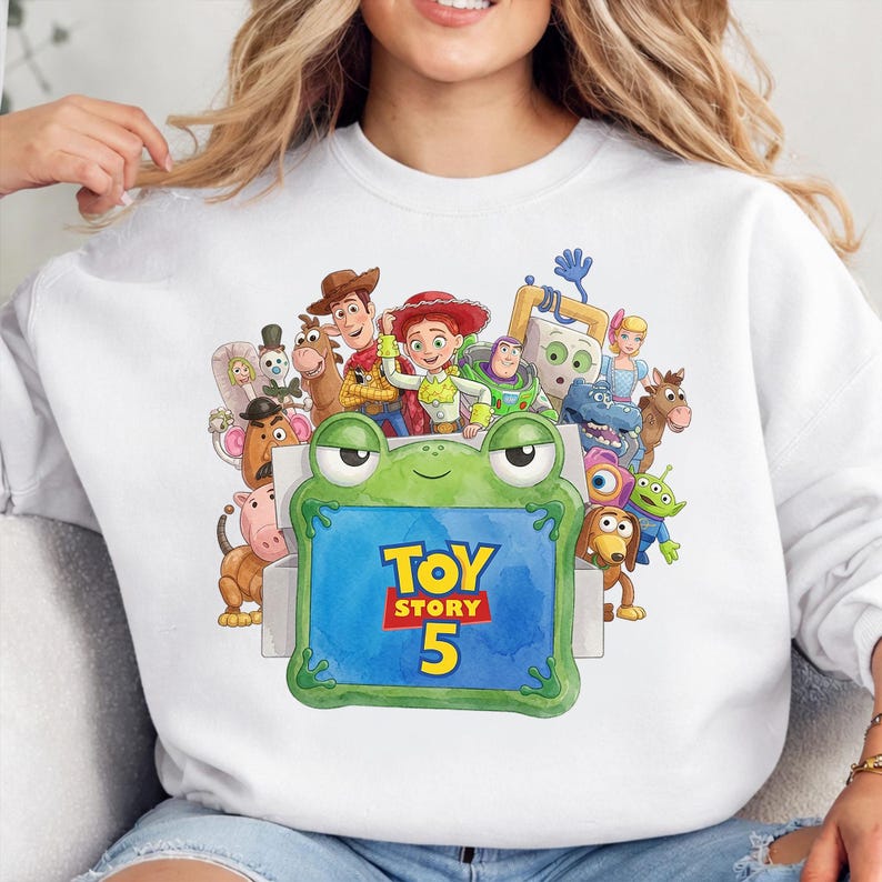 May include: A white crewneck sweatshirt featuring a colorful graphic print of various Toy Story characters surrounding a green frog-shaped frame. The center of the frame displays the blue text Toy Story 5 in a bold yellow and red font style.