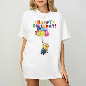 K&ouml;nnte beinhalten: Ein wei&szlig;es T-Shirt mit einer Grafik einer gelben Minion-Figur, die einen Strau&szlig; bunter Luftballons h&auml;lt. &Uuml;ber der Figur ist der Text "HAPPY BIRTHDAY" in bunten Buchstaben gedruckt, flankiert von zwei gelben Sternen auf jeder Seite.