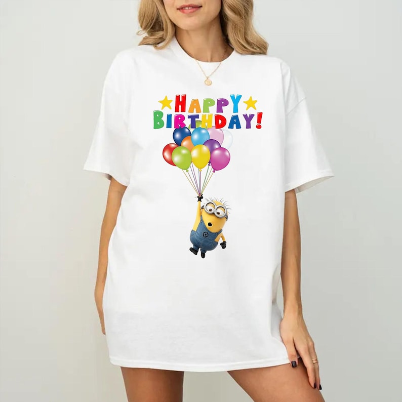 K&ouml;nnte beinhalten: Ein wei&szlig;es T-Shirt mit einer Grafik einer gelben Minion-Figur, die einen Strau&szlig; bunter Luftballons h&auml;lt. &Uuml;ber der Figur ist der Text "HAPPY BIRTHDAY" in bunten Buchstaben gedruckt, flankiert von zwei gelben Sternen auf jeder Seite.