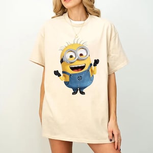 K&ouml;nnte beinhalten: Ein beiges Kurzarm-T-Shirt mit einem Grafikdruck eines gelben Minion-Charakters, der eine blaue Jeans-Latzhose, schwarze Handschuhe und eine silberne Schutzbrille tr&auml;gt. Die Figur ist mit einem fr&ouml;hlichen Ausdruck und erhobenen Armen dargestellt.