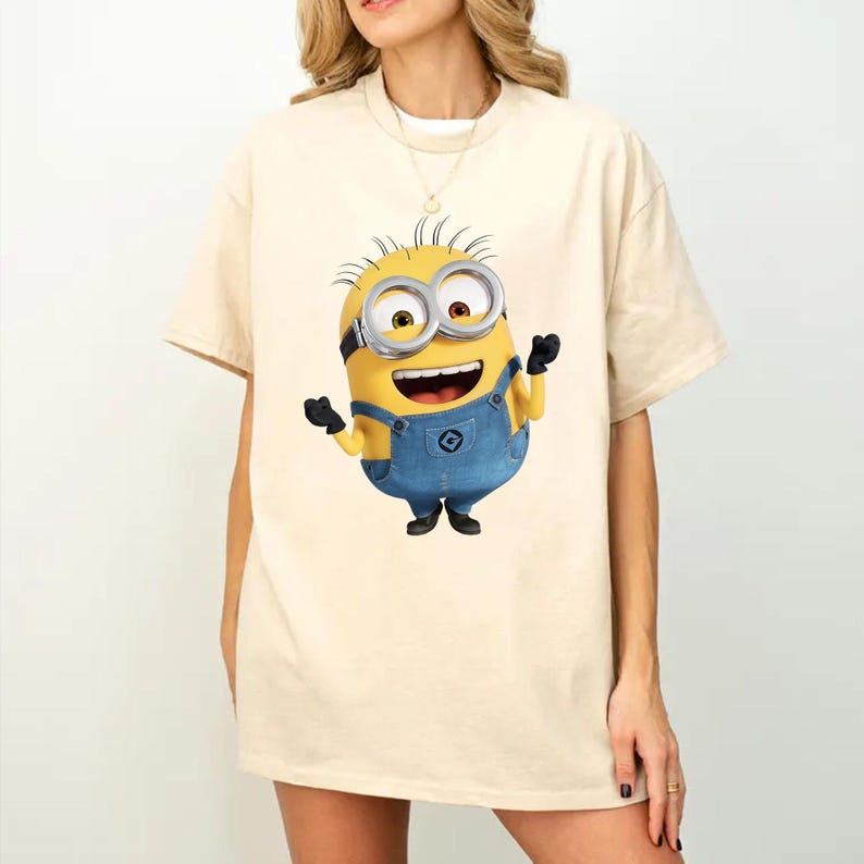 K&ouml;nnte beinhalten: Ein beiges Kurzarm-T-Shirt mit einem Grafikdruck eines gelben Minion-Charakters, der eine blaue Jeans-Latzhose, schwarze Handschuhe und eine silberne Schutzbrille tr&auml;gt. Die Figur ist mit einem fr&ouml;hlichen Ausdruck und erhobenen Armen dargestellt.