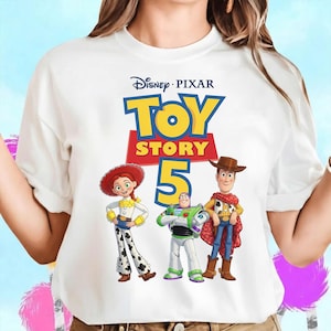 May include: A white short-sleeved t-shirt featuring a graphic print of the Disney Pixar Toy Story 5 logo with characters Jessie, Buzz Lightyear, and Woody standing below the text. The design includes the yellow and red logo with the number five.