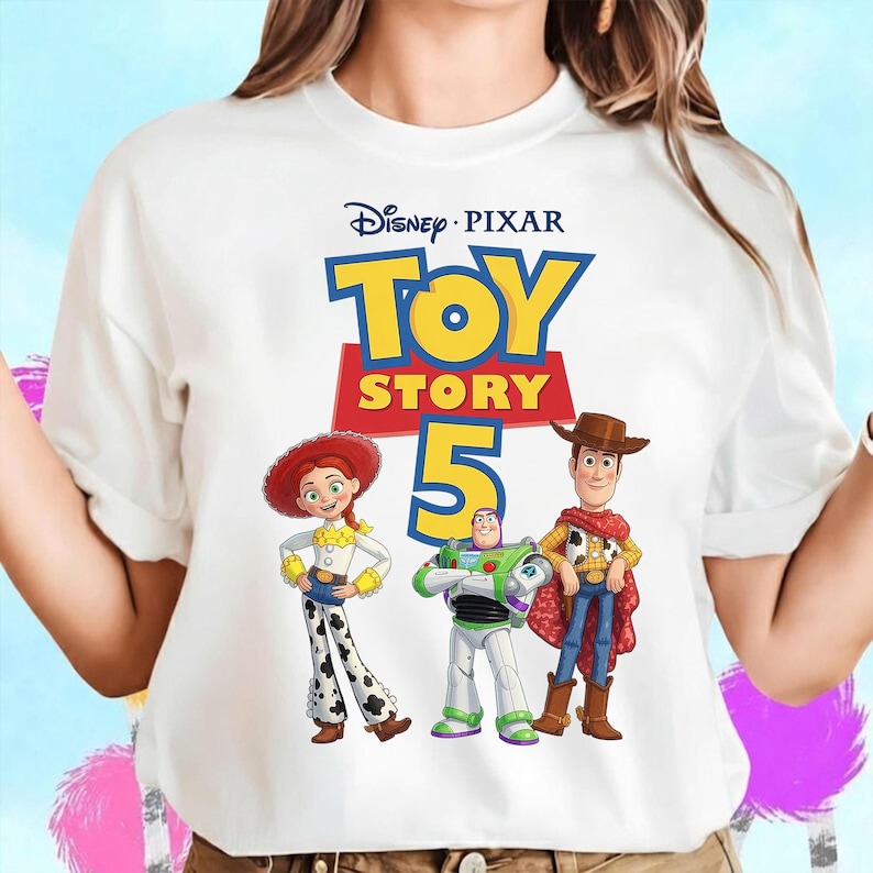 May include: A white short-sleeved t-shirt featuring a graphic print of the Disney Pixar Toy Story 5 logo with characters Jessie, Buzz Lightyear, and Woody standing below the text. The design includes the yellow and red logo with the number five.