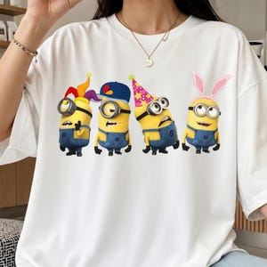 K&ouml;nnte beinhalten: Ein wei&szlig;es T-Shirt mit kurzen &Auml;rmeln und einem Grafikdruck von vier gelben Minion-Figuren. Die Figuren tragen blaue Jeans-Latzhosen und verschiedene Accessoires, darunter eine Narrenkappe, eine blaue Baseballkappe, einen rosa Partyhut und Hasenohren.