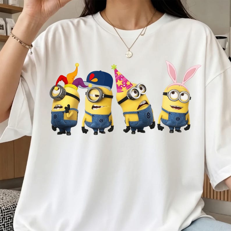 K&ouml;nnte beinhalten: Ein wei&szlig;es T-Shirt mit kurzen &Auml;rmeln und einem Grafikdruck von vier gelben Minion-Figuren. Die Figuren tragen blaue Jeans-Latzhosen und verschiedene Accessoires, darunter eine Narrenkappe, eine blaue Baseballkappe, einen rosa Partyhut und Hasenohren.