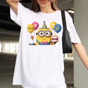 K&ouml;nnte beinhalten: Ein wei&szlig;es T-Shirt mit kurzen &Auml;rmeln und einer Grafik eines gelben Minions, der einen blau-gelben Partyhut und eine rosa Fliege tr&auml;gt. Der Minion h&auml;lt einen Geburtstagskuchen sowie mehrere bunte Luftballons in Gelb, Rosa und Blau in den H&auml;nden.