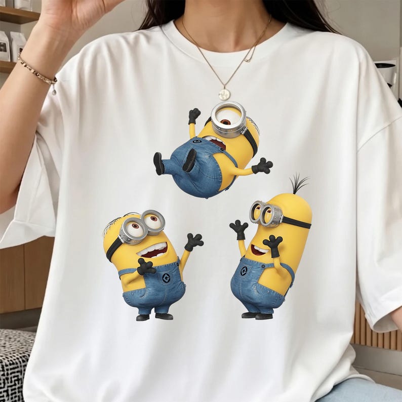 K&ouml;nnte beinhalten: Ein wei&szlig;es T-Shirt mit kurzen &Auml;rmeln und einem Grafikdruck von drei gelben Minion-Figuren, die blaue Jeans-Latzhosen und silberne Schutzbrillen tragen. Die Figuren sind in verschiedenen Posen dargestellt, wobei eine &uuml;ber den anderen schwebt.