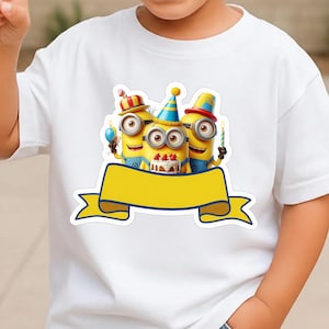 K&ouml;nnte beinhalten: Ein wei&szlig;es Kurzarm-T-Shirt mit einer Grafik von drei gelben Minion-Figuren, die bunte Partyh&uuml;te tragen und eine Geburtstagstorte mit Kerzen halten, platziert &uuml;ber einem gro&szlig;en, leeren gelben Banner mit dunkelblauem Rand.