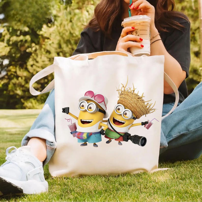 K&ouml;nnte beinhalten: Eine wei&szlig;e Stofftasche mit einem Grafikdruck von zwei gelben Minion-Figuren. Ein Minion tr&auml;gt einen wei&szlig;en Hut und blaue Shorts mit einer regenbogenfarbenen Bauchtasche, der andere einen Strohhut und gr&uuml;ne Shorts und h&auml;lt eine Kamera.
