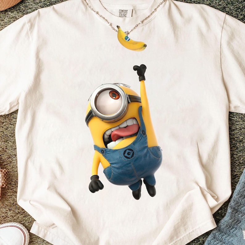 K&ouml;nnte beinhalten: Ein cremefarbenes T-Shirt mit einer Grafik eines gelben, ein&auml;ugigen Minion-Charakters, der eine blaue Jeanslatzhose mit einem schwarzen Logo tr&auml;gt. Der Charakter hat den Mund offen, die Zunge herausgestreckt und einen Arm erhoben. Eine silberne Kette mit Bananenanh&auml;nger.