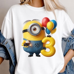 K&ouml;nnte beinhalten: Ein wei&szlig;es T-Shirt mit einer Grafik eines gelben, ein&auml;ugigen Minion-Charakters in blauer Latzhose. Die Figur h&auml;lt einen kleinen Cupcake mit einer brennenden Kerze und steht neben einem gro&szlig;en gelben Luftballon in Form einer Drei sowie einer Ansammlung roter, gelber und blauer Luftballons.