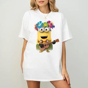 K&ouml;nnte beinhalten: Ein wei&szlig;es T-Shirt mit einer Grafik eines gelben Minion-Charakters, der eine bunte Blumenkette und einen gr&uuml;nen Bastrock tr&auml;gt. Der Minion spielt eine braune h&ouml;lzerne Ukulele und hat ein breites L&auml;cheln. Das Motiv ist mittig platziert.