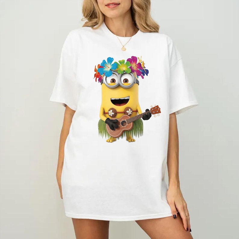 K&ouml;nnte beinhalten: Ein wei&szlig;es T-Shirt mit einer Grafik eines gelben Minion-Charakters, der eine bunte Blumenkette und einen gr&uuml;nen Bastrock tr&auml;gt. Der Minion spielt eine braune h&ouml;lzerne Ukulele und hat ein breites L&auml;cheln. Das Motiv ist mittig platziert.