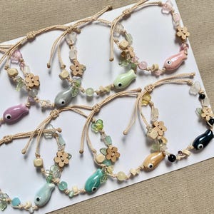 May include: A collection of handmade bracelets featuring colorful fish charms, wooden flower beads, and various gemstone beads. The bracelets are strung on beige cords and are displayed on a white surface.