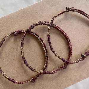 May include: Three circular bracelets crafted from twisted cord in deep burgundy and metallic gold tones. Each bracelet features a continuous braided pattern with alternating sections of dark red and shimmering gold thread, creating a textured look.