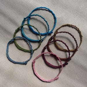 May include: A collection of seven thin, braided cord bracelets in various colors including light blue, dark blue, olive green, tan, dark brown, deep purple, and light pink. Each bracelet features a simple knotted closure and a woven texture.