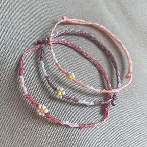 May include: Three thin, braided cord bracelets in shades of pink, purple, and white. Each bracelet features a small, delicate daisy-shaped bead with white petals and a yellow center, integrated into the woven cord design with adjustable knots.