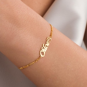 May include: A gold-toned chain bracelet featuring a personalized nameplate with the script text Chloe followed by a small paw print cutout. The delicate metal chain connects to each side of the nameplate, creating a continuous link design.