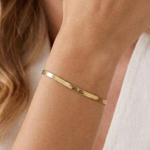 May include: A thin, polished gold-tone herringbone chain bracelet with a flat, flexible link design that rests smoothly against the wrist. The bracelet features a reflective metallic surface and a secure clasp, showcasing a sleek and uniform appearance.