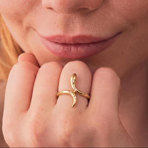 May include: A gold-coloured ring shaped like a snake, with a curved body and a head at the top. The ring is decorated with small, clear gemstones. The ring is on a finger.