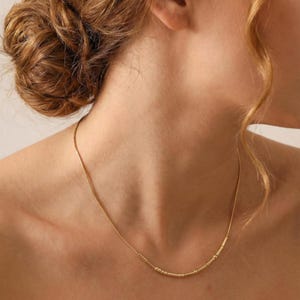 May include: A delicate gold necklace with a thin chain and a series of small, evenly spaced cylindrical and rectangular beads. The necklace is worn around the neck, and the design is simple and elegant.