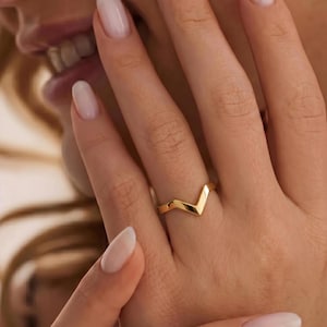 May include: A gold-colored ring with a V-shaped design. The ring is worn on a finger, and the design is a simple, modern style. The ring is likely made of gold or a gold-colored metal.
