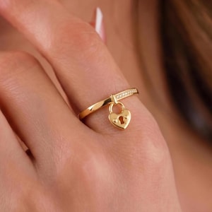 May include: A gold-colored ring featuring a heart-shaped padlock pendant. The ring band is a simple, twisted design, with a section of the band adorned with small, clear stones. The padlock has a keyhole detail.