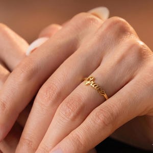 Pode incluir: Um anel dourado com o nome "Grace" em fonte cursiva. O anel é usado num dedo, mostrando o design delicado e a inscrição personalizada. O anel é uma joia.