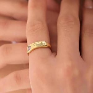May include: A gold-colored ring with the name "Taylor" engraved on the band. The ring features a small, round, light-colored stone. The ring is worn on a finger, with the hand in soft focus.